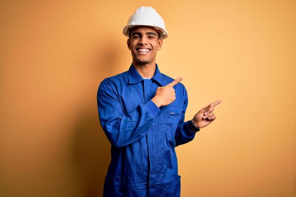 młody przystojny afroamerykański pracownik ubrany w niebieski mundur i kask ochronny uśmiecha się i patrzy na kamerę skierowaną dwoma rękami i palcami na bok. - isolated professional occupation engineer human face zdjęcia i obrazy z banku zdjęć