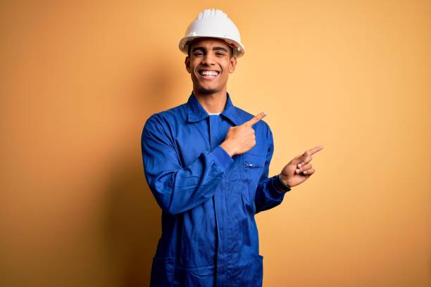 jovem bonito trabalhador afro-americano vestindo uniforme azul e capacete de segurança sorrindo e olhando para a câmera apontando com duas mãos e dedos para o lado. - vestuário para proteção - fotografias e filmes do acervo