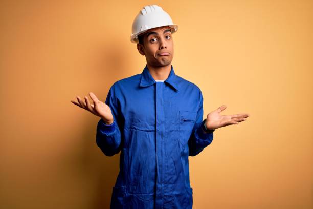 istock Young handsome african american worker man wearing blue uniform and security helmet clueless and confused expression with arms and hands raised. Doubt concept. 1214212038