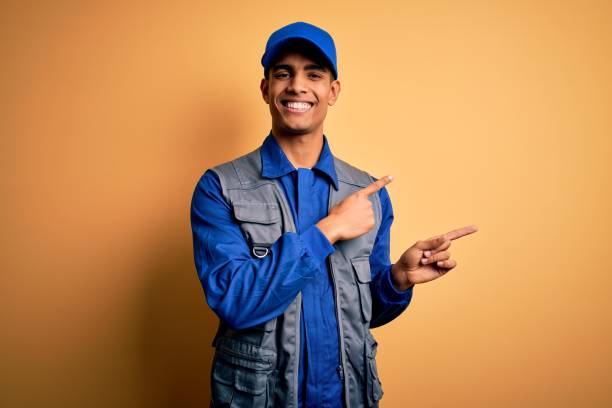 Young handsome african american handyman wearing worker uniform and cap smiling and looking at the camera pointing with two hands and fingers to the side. Young handsome african american handyman wearing worker uniform and cap smiling and looking at the camera pointing with two hands and fingers to the side. sign human hand pointing manual worker stock pictures, royalty-free photos & images
