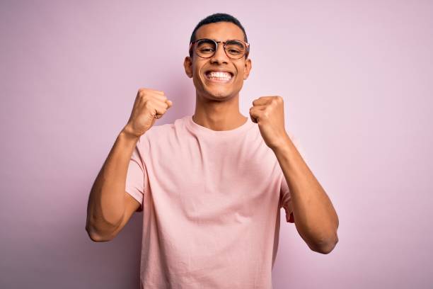 bel uomo afro-americano che indossa maglietta casual e occhiali su sfondo rosa eccitato per il successo con le braccia alzate e gli occhi chiusi che celebrano la vittoria sorridendo. concetto di vincitore. - afro americano foto e immagini stock