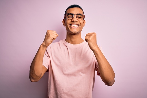 Handsome african american man wearing casual t-shirt and glasses over pink background excited for success with arms raised and eyes closed celebrating victory smiling. Winner concept.