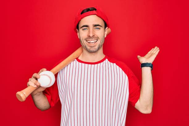 jeune homme sportif beau avec des yeux bleus jouant au base-ball portant la batte et la bille très heureuse et excitée, expression de gagnant célébrant la victoire criant avec le grand sourire et les mains soulevées - men baseball baseball cap baseball bat photos et images de collection