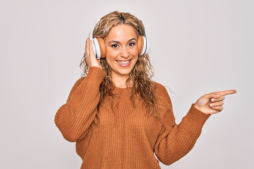 Young beautiful blonde woman listening to music using headphones over white background Smiling happy pointing with hand and finger to the side