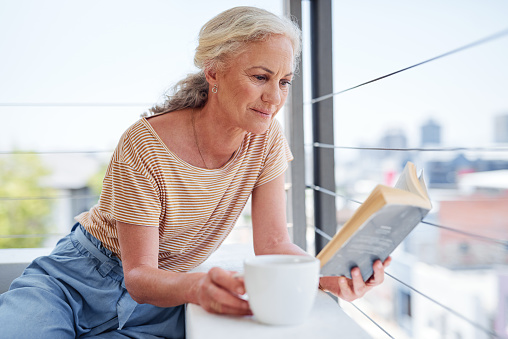 Cropped shot of an attractive senior woman enjoying a cup of coffee while reading a book outside on her balcony at home