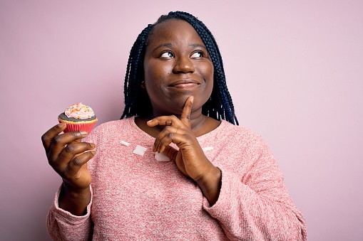 Young african american plus size woman with braids eating sweet chocolate cupcake serious face thinking about question, very confused idea