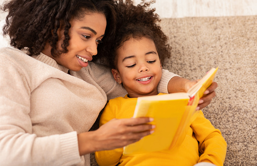 Cute Black Mother And Little Daughter Reading Book Together Sitting On Couch At Home. Bedtime Story Concept