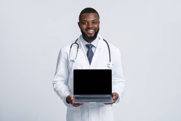 istock Black cheerful doc in white robe holding blank laptop screen 1214222349