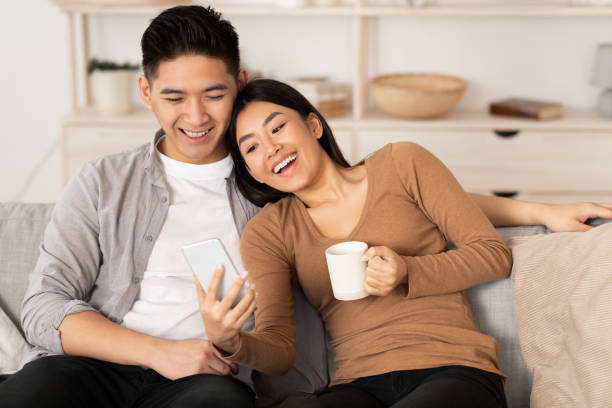Asian couple resting on couch, using phone Spending Time Together. Asian family using smart phone sitting on couch at home, empty space happy filipino family stock pictures, royalty-free photos & images