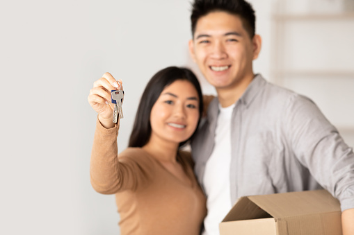 Homeowners. Smiling chinese couple showing key to camera, man holding box, selective focus, copy space