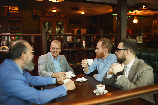 Group of businessmen in suits sitting at the table with cups of coffee and talking to each other during meeting in cafe