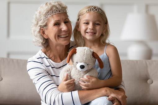 Family portrait of overjoyed senior grandmother and cute little granddaughter wearing crowns play funny game together in living room, smiling mature granny and small preschooler grandchild have fun