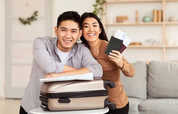 Travel Concept. Chinese Couple Holding Boarding Pass And Passports, Ready For Vacation Abroad, Sitting With Suitcase On Sofa At Home