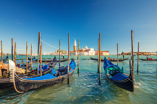 Gondolas moored docked on water in Venice. Gondoliers sailing San Marco basin waterway. San Giorgio Maggiore island with Campanile San Giorgio in Venetian Lagoon, blue clear sky, Veneto Region, Italy