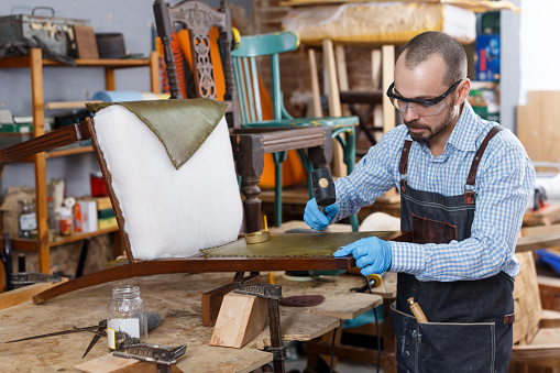 Positive craftsman engaged in repair of vintage furniture, reupholstering chair in workshop