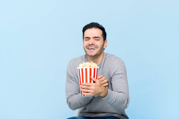 istock Young man holding popcorn laughing while watching movie isolated on light blue background 1214232711