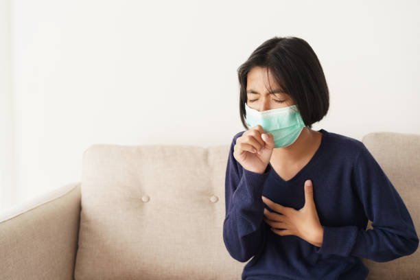 Asian girl symptom cough and are protective with medical mask while sitting on sofa, Asia child wearing a protection mask epidemic of flu or covid-19 in living room at home. Health and illness concept stock photo