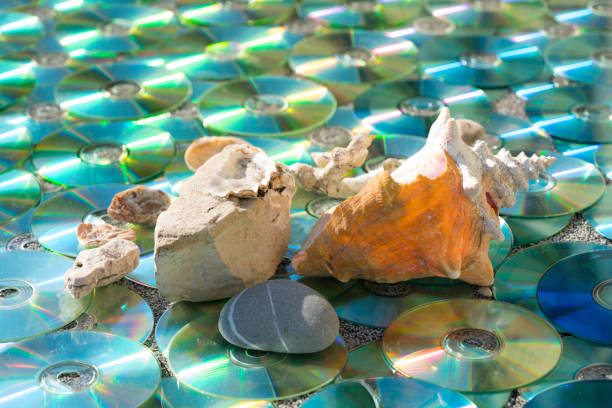 shells and rocks with sun reflection as water of multicolored, old technology, waste CD/DVD as decoration for pattern, textured background.