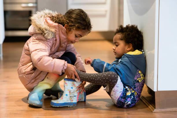 Sisterly Love Mixed race sisters, dressed in their warm coats, getting ready to go outside and play. The older sibling is helping the younger sister put on her wellington boots. affectionate stock pictures, royalty-free photos & images