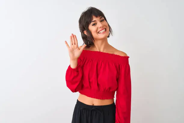 Photo of Young beautiful woman wearing red summer t-shirt standing over isolated white background Waiving saying hello happy and smiling, friendly welcome gesture