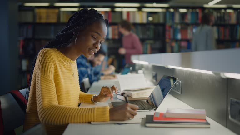 University Library: Gifted Beautiful Black Girl Sitting at the Desk, Uses Laptop, Writes Notes for the Paper, Essay, Study for Class Assignment. Diverse Group of Students Learning, Studying for Exams.