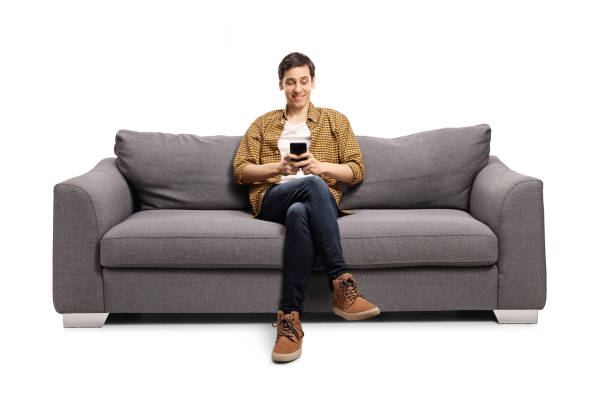 glücklicher junger mann sitzt auf einem grauen sofa und tippt auf ein handy - sitzen stock-fotos und bilder