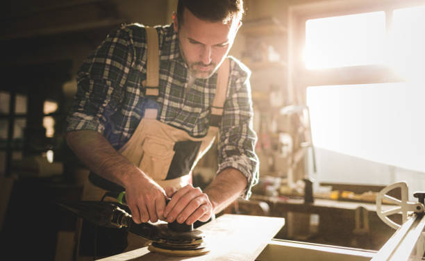 istock Carpenter at workshop polishes wooden board with a electric orbital sander 1214255355