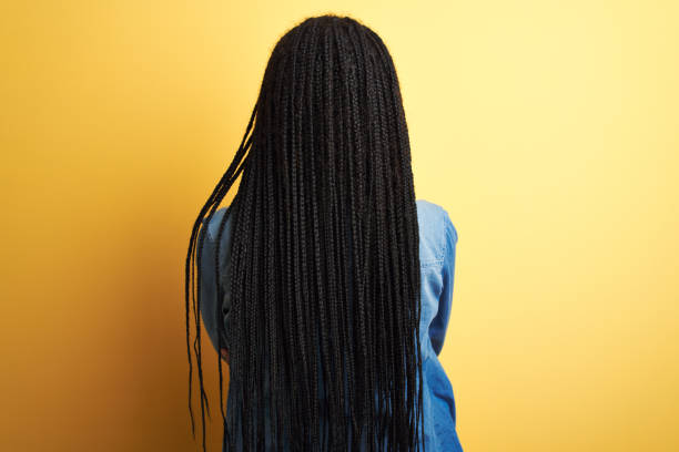 孤立した黄色の背景の上に立っているデニムシャツを着た若いアフリカ系アメリカ人女性は、腕を組んで後ろを見て立っています - 編み込みヘア ストックフォトと画像