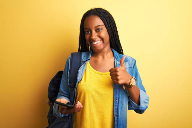 istock African american student woman using smartphone standing over isolated yellow background happy with big smile doing ok sign, thumb up with fingers, excellent sign 1214255655