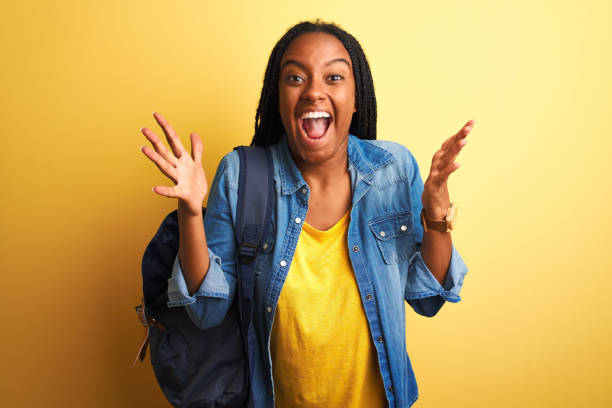 istock African american student woman wearing backpack standing over isolated yellow background celebrating crazy and amazed for success with arms raised and open eyes screaming excited. Winner concept 1214255721