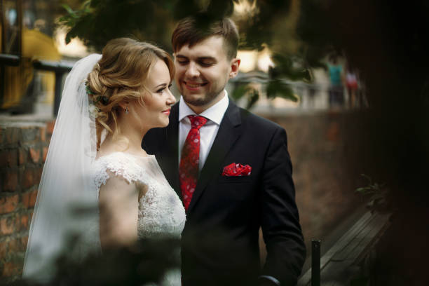 couples heureux de jeune marié étreignant à l’extérieur dans la vieille rue italienne, belle mariée blonde dans la robe de mariée blanche de dentelle et beau marié posant près de la nature - veil bride lace married photos et images de collection