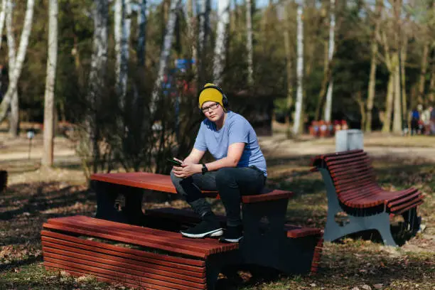 March 18, 2020 - Kabaty, Poland:Sun and shadows in the picnic area in a park with a teenage Caucasian boy in blue shirt sitting on top of the table, holding e-reader, listening to music on his headphones, looking at camera, being healthy