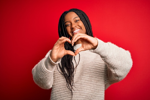Young african american woman wearing casual winter sweater over red isolated background smiling in love doing heart symbol shape with hands. Romantic concept.
