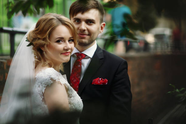 couples heureux de jeune marié étreignant à l’extérieur dans la vieille rue italienne, belle mariée blonde dans la robe de mariée blanche de dentelle et beau marié posant près de la nature - veil bride lace married photos et images de collection