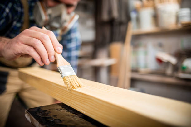 istock Carpenter with mask applies paint using paintbrush in carpentry workshop 1214260102