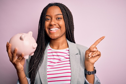 Young african american business woman saving money on piggy bank over isolated background very happy pointing with hand and finger to the side