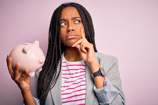 Young african american business woman saving money on piggy bank over isolated background serious face thinking about question, very confused idea