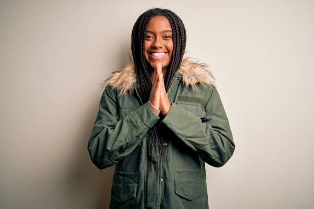 молодая афроамериканка в зимнем пальто парка на изолированном фоне молится с руками и уверенно просит прощения, уверенно улыбаясь. - braided women african descent winter стоковые фото и изображения