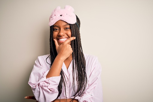 Young african american woman wearing pink pajama and sleep mask over isolated background looking confident at the camera smiling with crossed arms and hand raised on chin. Thinking positive.