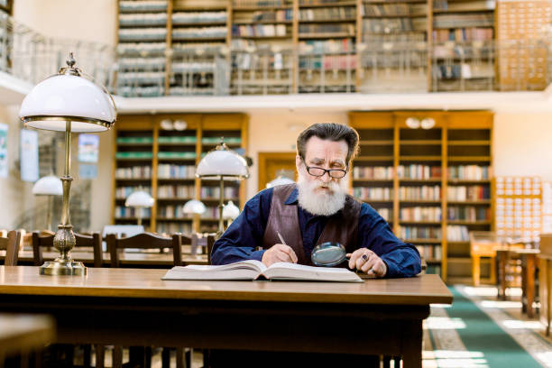 istock Senior man working in old library, sitting at the table with books and magnifying glass, making notes. Student old man, education in library, old shelves with books on the background 1214267465