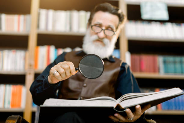 bell'uomo barbuto anziano sorridente, bibliotecario o professore, in biblioteca, seduto sullo sfondo delle librerie, tenendo la lente d'ingrandimento e leggendo libro. concentrati sul vetro e sul libro - archivista foto e immagini stock