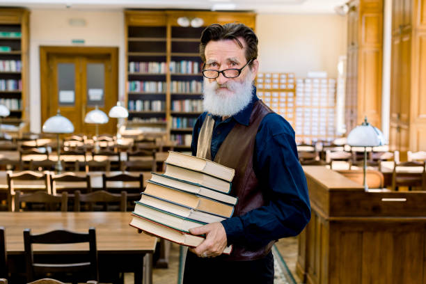 ritratto di sicuro elegante bibliotecario del professore universitario insegnante uomo, indossando abiti eleganti, felice di condividere conoscenze, tenendo una pila di libri diversi, in piedi nella biblioteca vintage al chiuso. - professor library librarian university foto e immagini stock