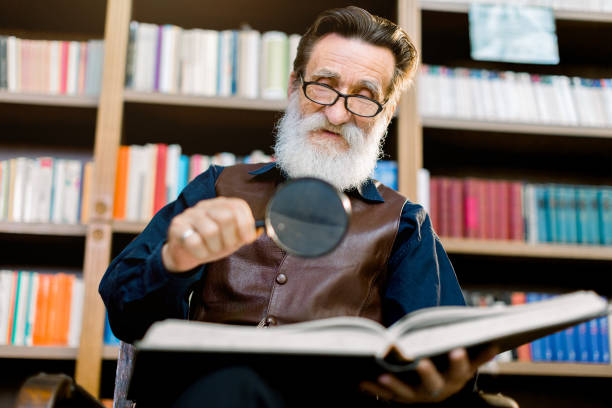 bel uomo barbuto anziano pensile, bibliotecario o professore, in biblioteca, seduto sullo sfondo delle librerie, tenendo la lente d'ingrandimento e leggendo libro - archivista foto e immagini stock