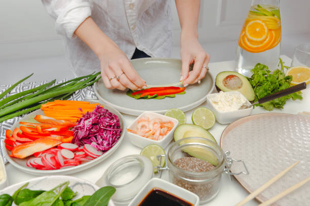 istock Woman's hands putting red pepper and cucumbers on rice paper. Table with spring rolls ingredients and water with oranges. 1214272905