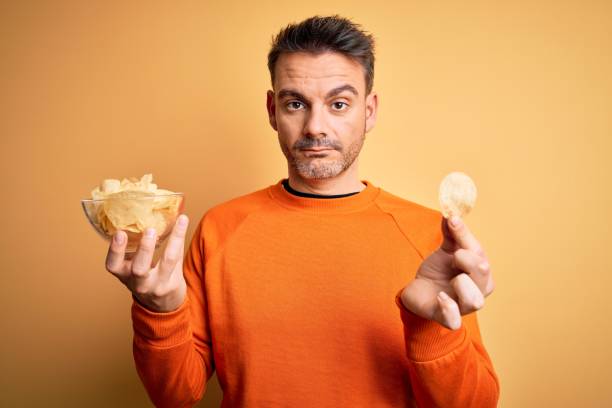 istock Young handsome man holding bowl with potatoes chips over isolated yellow background with a confident expression on smart face thinking serious 1214274934