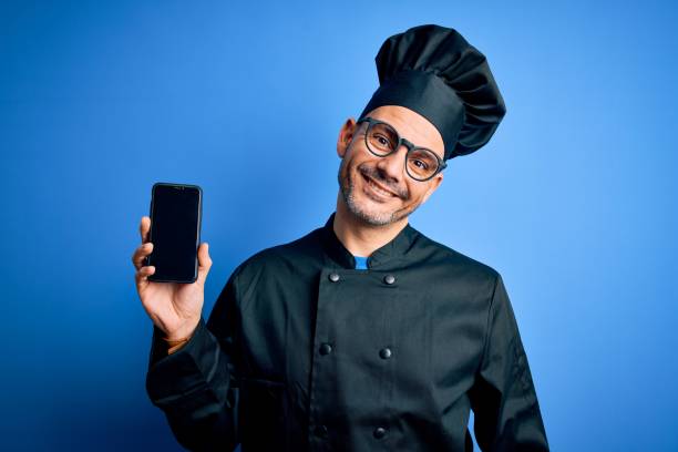 istock Young handsome chef man wearing uniform and hat holding smartphone over blue background with a happy face standing and smiling with a confident smile showing teeth 1214276016