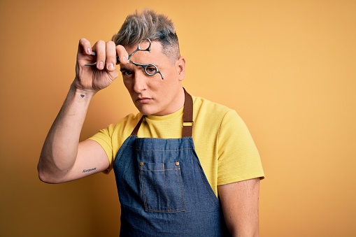 Young handsome blonde hairdresser man wearing apron holding scissors standing over isolated yellow background