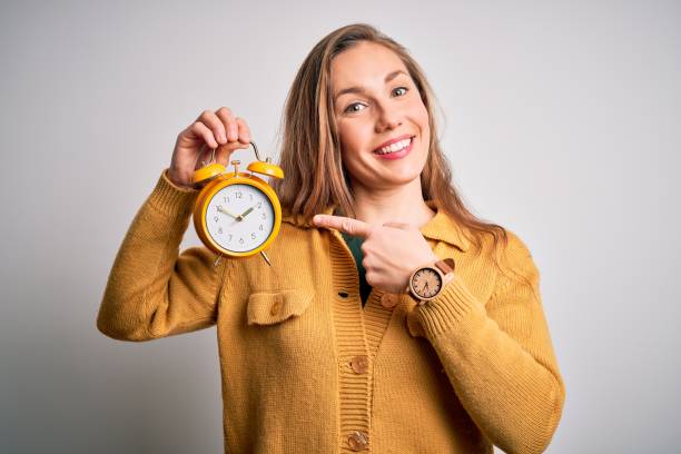 young beautiful blonde woman holding alarm clock standing over isolated white background very happy pointing with hand and finger - clock face fotos imagens e fotografias de stock