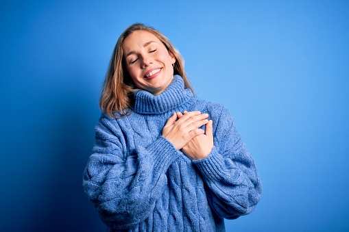 Young beautiful blonde woman wearing casual turtleneck sweater over blue background smiling with hands on chest with closed eyes and grateful gesture on face. Health concept.