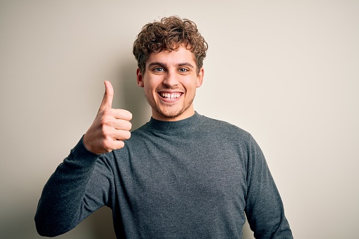 Young blond handsome man with curly hair wearing casual sweater over white background doing happy thumbs up gesture with hand. Approving expression looking at the camera showing success.